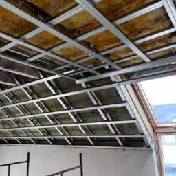 Sufit podwieszany z izolacją z wełny mineralnej i metalową konstrukcją, widok z dołu. Fragment rusztowania i okno dachowe w tle. Prace wykończeniowe.