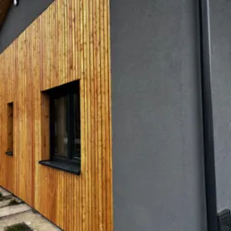 Elewacja domu z drewnianą okładziną i szarym tynkiem. Widoczne okna z czarnymi ramami i rynna. Nowoczesny design z naturalnymi materiałami.