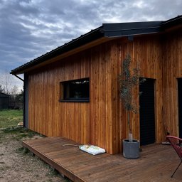 Usługi remontowo budowlane Roman Kapituła - Nowoczesny dom z drewnianą elewacją i tarasem w ciepłym odcieniu brązu. Minimalistyczne okno, czarne elementy wykończenia i dekoracyjne drzewko w donicy.