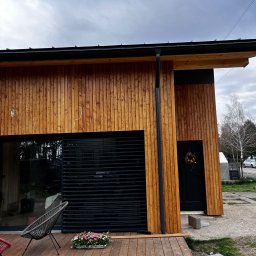 Usługi remontowo budowlane Roman Kapituła - Nowoczesny dom z drewnianą elewacją, czarnymi żaluzjami i drewnianym tarasem. Widoczny fragment ogrodu i dekoracja na drzwiach wejściowych.