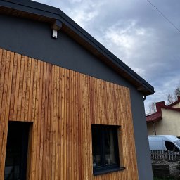 Usługi remontowo budowlane Roman Kapituła - Elewacja domu z drewnianą okładziną i szarą strukturą, widok z dołu, z oknami w czarnych ramach. W tle budynki i błękitne niebo.