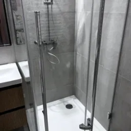 Nowoczesna kabina prysznicowa z chromowanymi detalami i szarymi płytkami. Widoczna umywalka i odbicie osoby w szkle. Minimalistyczny design.