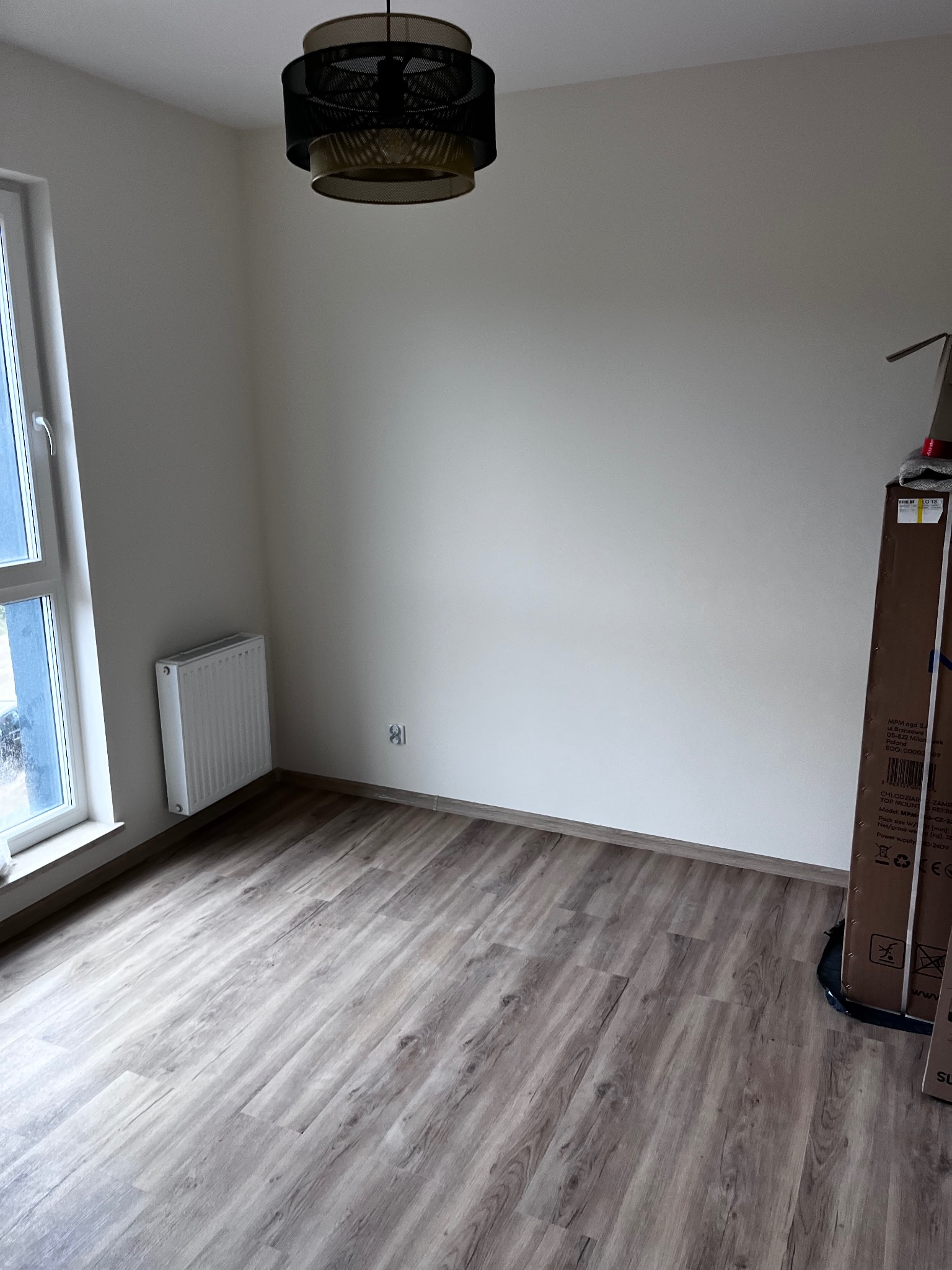 Pokój po remoncie: panele podłogowe w odcieniach szarości, białe ściany, okno z grzejnikiem, lampa sufitowa. Częściowo widoczny karton z urządzeniem AGD.