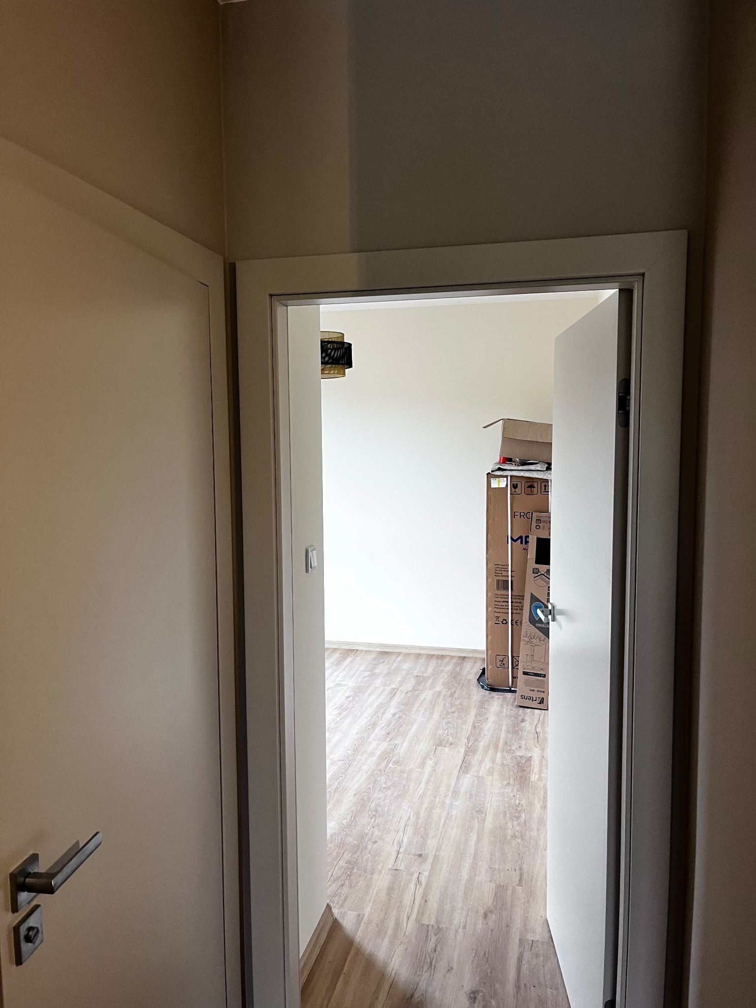 Otwarta, beżowa, wewnętrzna rama drzwiowa z metalową klamką, przez którą widać pokój z panelami i kartonami. Widok z perspektywy korytarza.