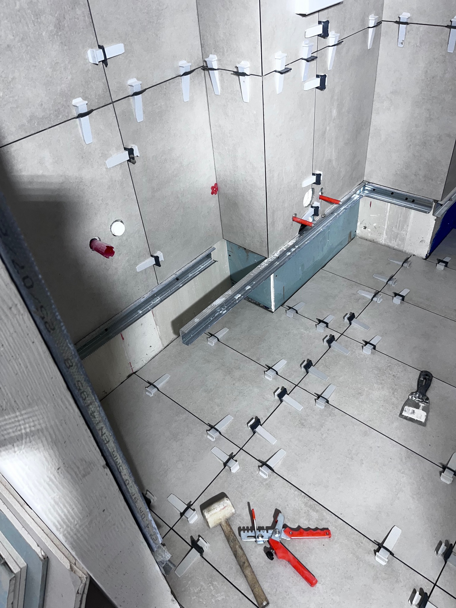 Jasnoszare płytki w trakcie układania z użyciem systemu poziomowania. Widoczne narzędzia: młotek, poziomica, kielnia. Fragment zabudowy z profili.