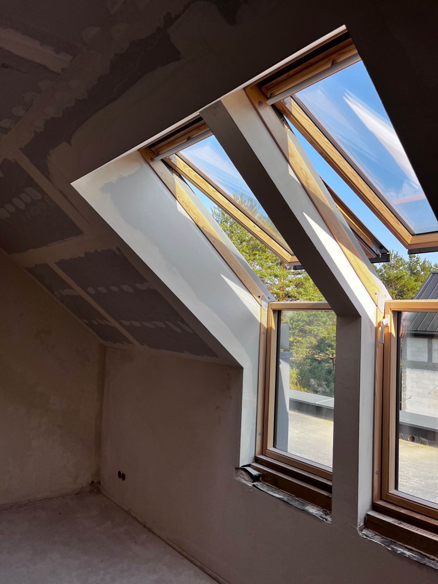 Pokój na poddaszu w trakcie wykończenia, widoczne okna dachowe z drewnianymi ramami, ściany przygotowane do malowania, sufit z płyt kartonowo-gipsowych.