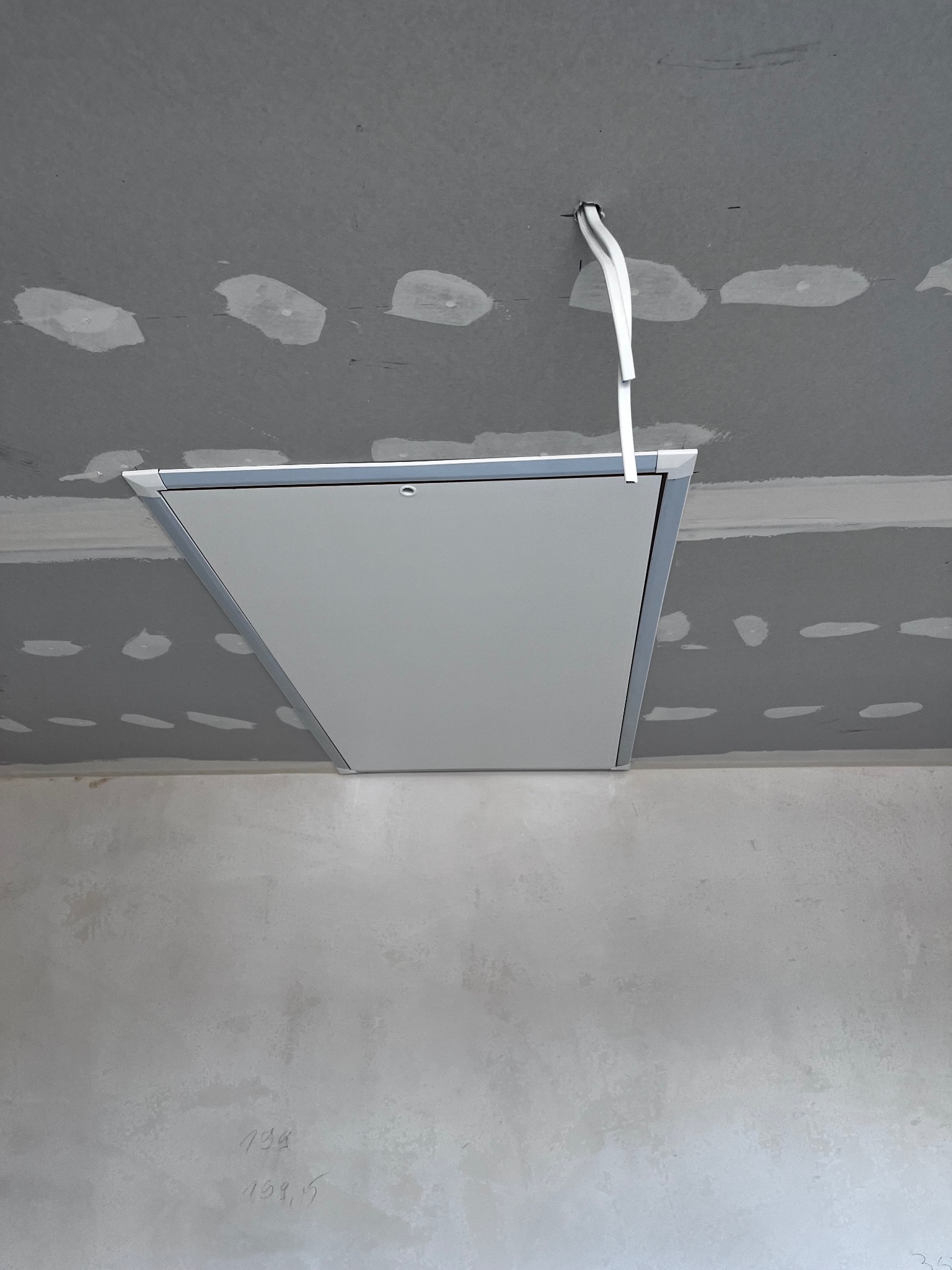 Zamknięta klapa rewizyjna w szarej płycie kartonowo-gipsowej z widocznymi przewodami i śladami szpachlowania sufitu oraz surową ścianą w tle.