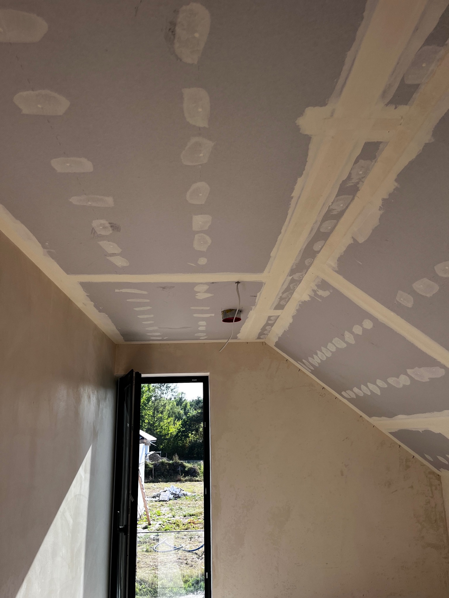Płyty karton-gips na suficie poddasza, wstępnie zaszpachlowane łączenia, widok na okno i fragment ściany, etap prac wykończeniowych.