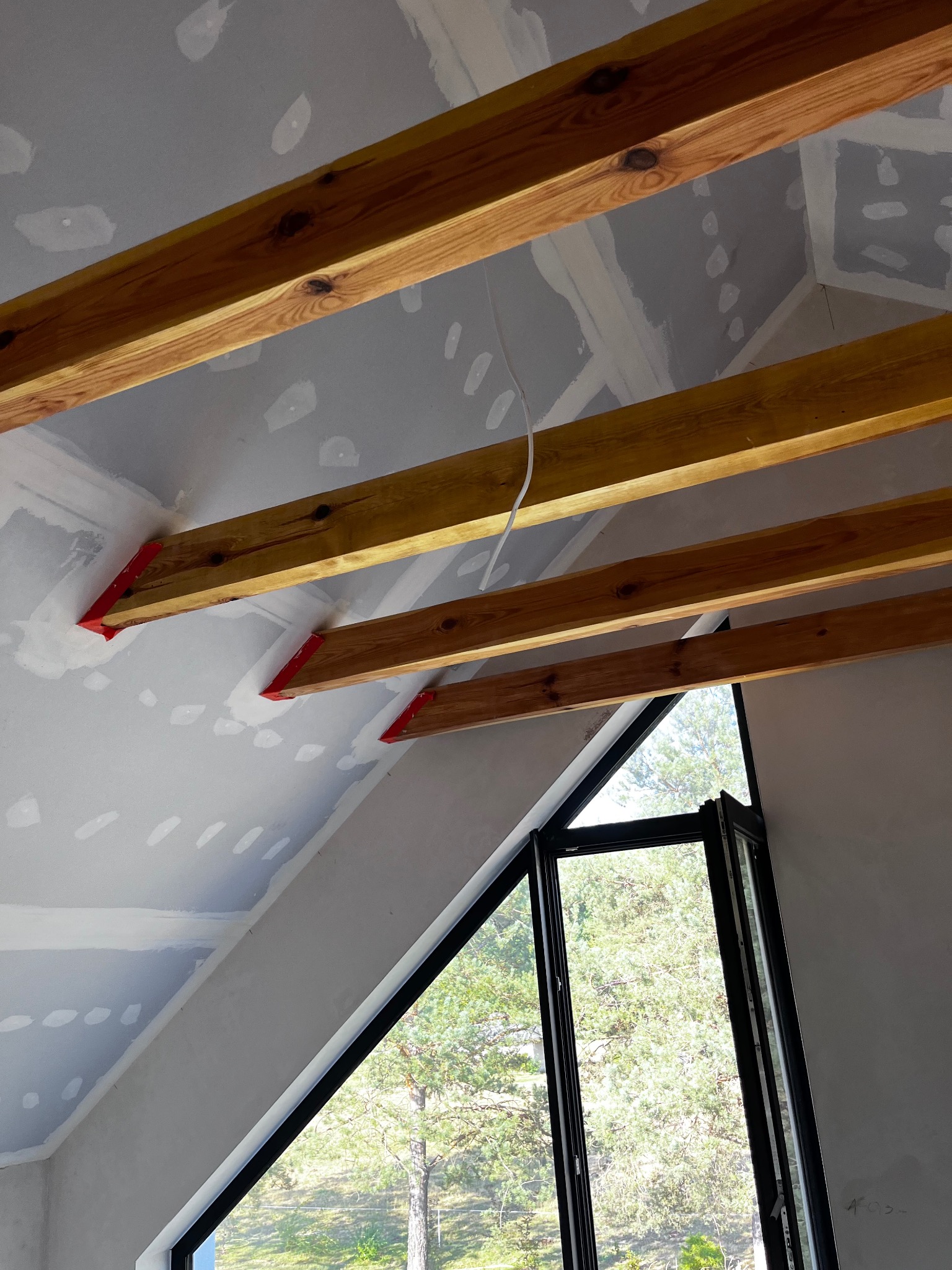 Skośny sufit z płyt karton-gips, drewniane belki z czerwonymi wspornikami, widok na zieleń przez okno. Etap prac wykończeniowych, szpachlowanie sufitu.