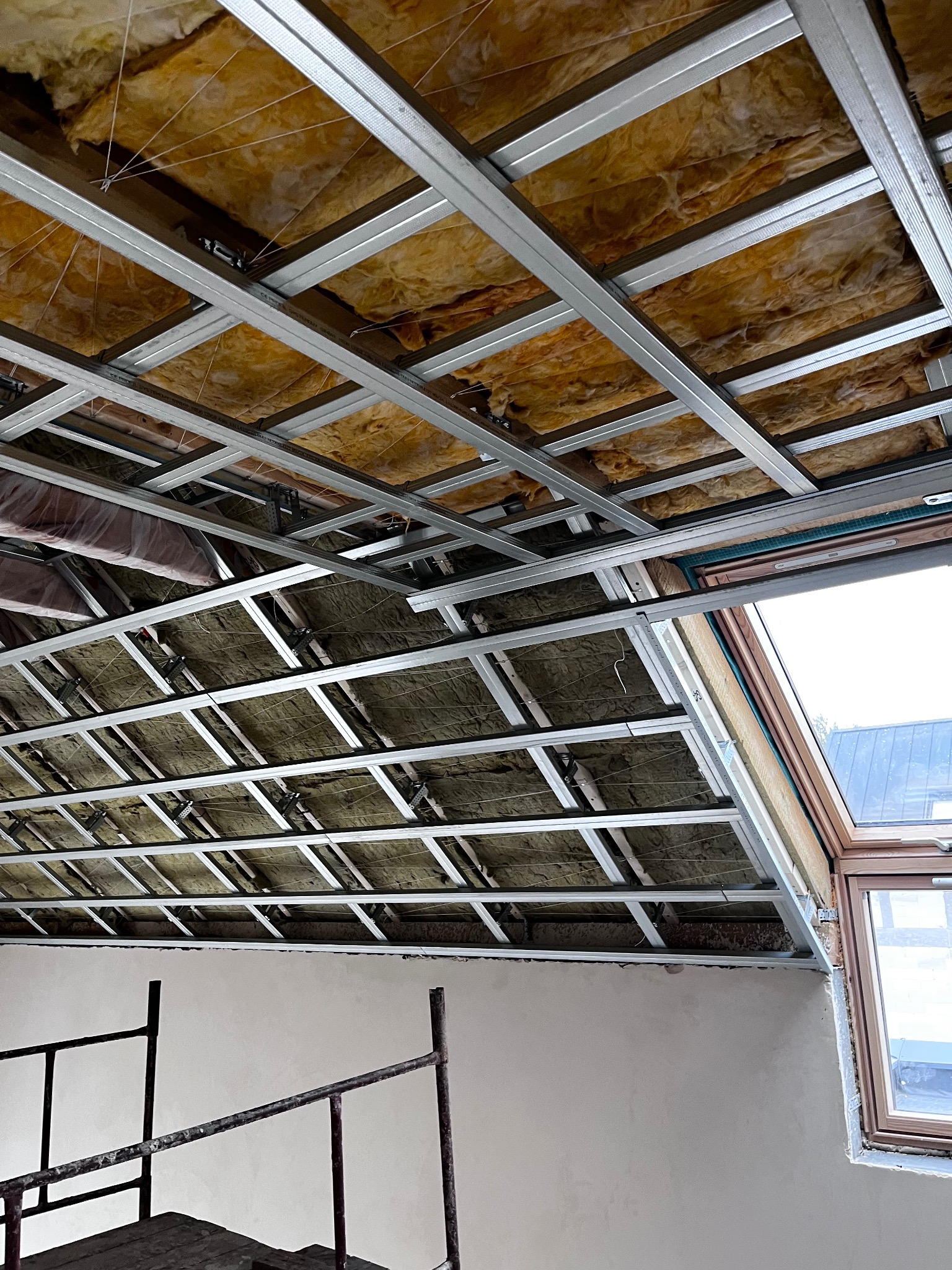 Sufit podwieszany z izolacją z wełny mineralnej i metalową konstrukcją, widok z dołu. Fragment rusztowania i okno dachowe w tle. Prace wykończeniowe.