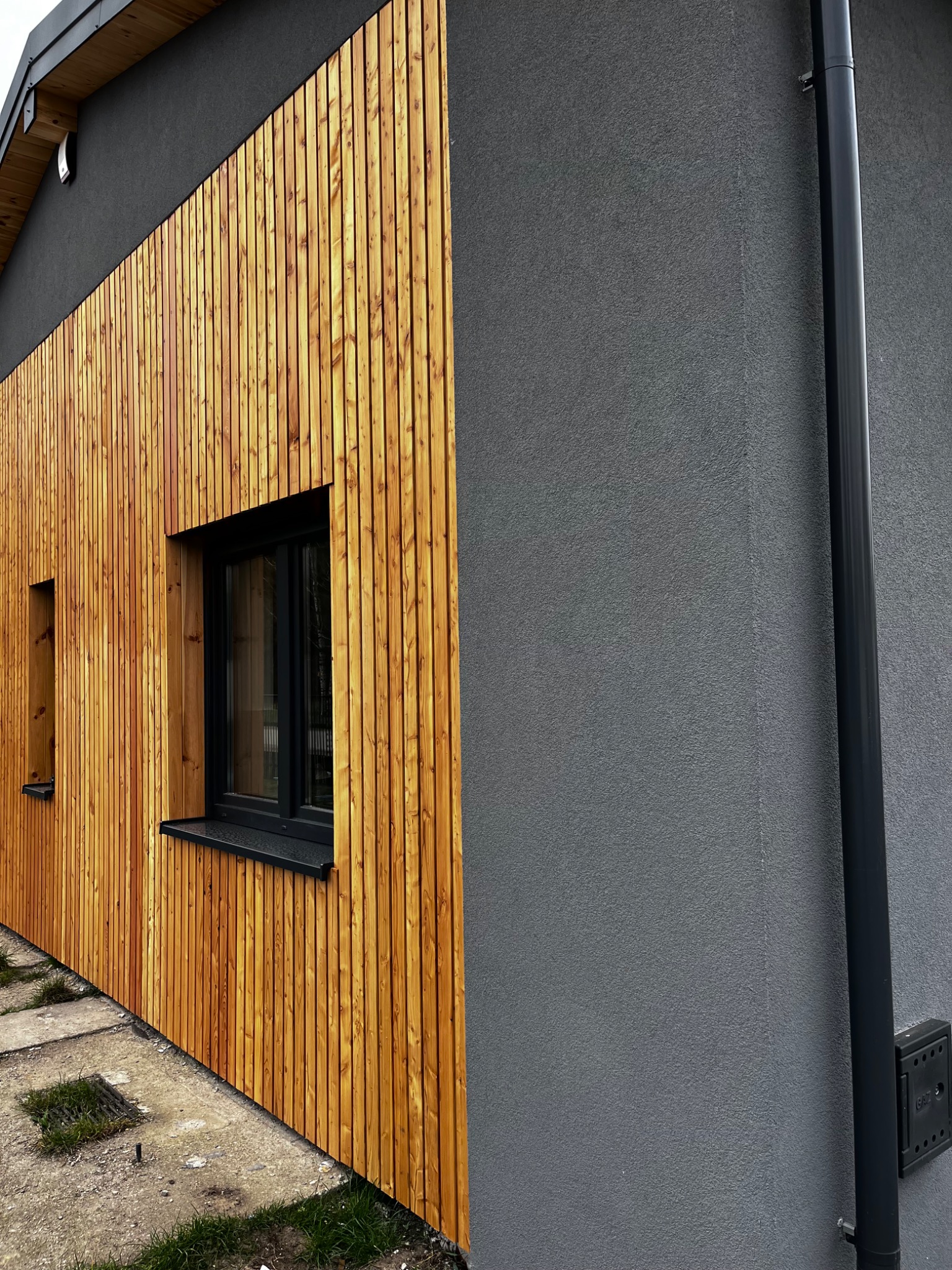 Elewacja domu z drewnianą okładziną i szarym tynkiem. Widoczne okna z czarnymi ramami i rynna. Nowoczesny design z naturalnymi materiałami.