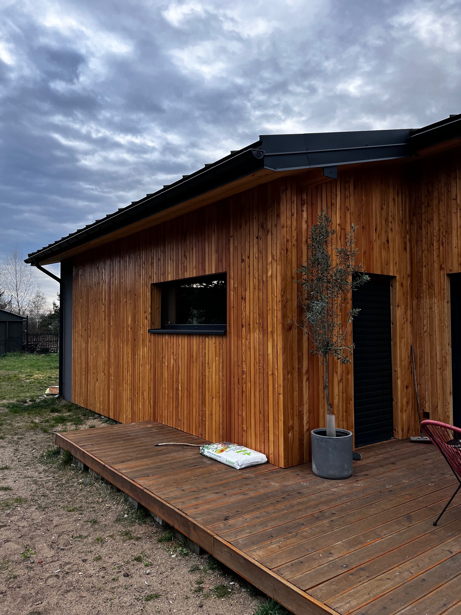 Nowoczesny dom z drewnianą elewacją i tarasem w ciepłym odcieniu brązu. Minimalistyczne okno, czarne elementy wykończenia i dekoracyjne drzewko w donicy.