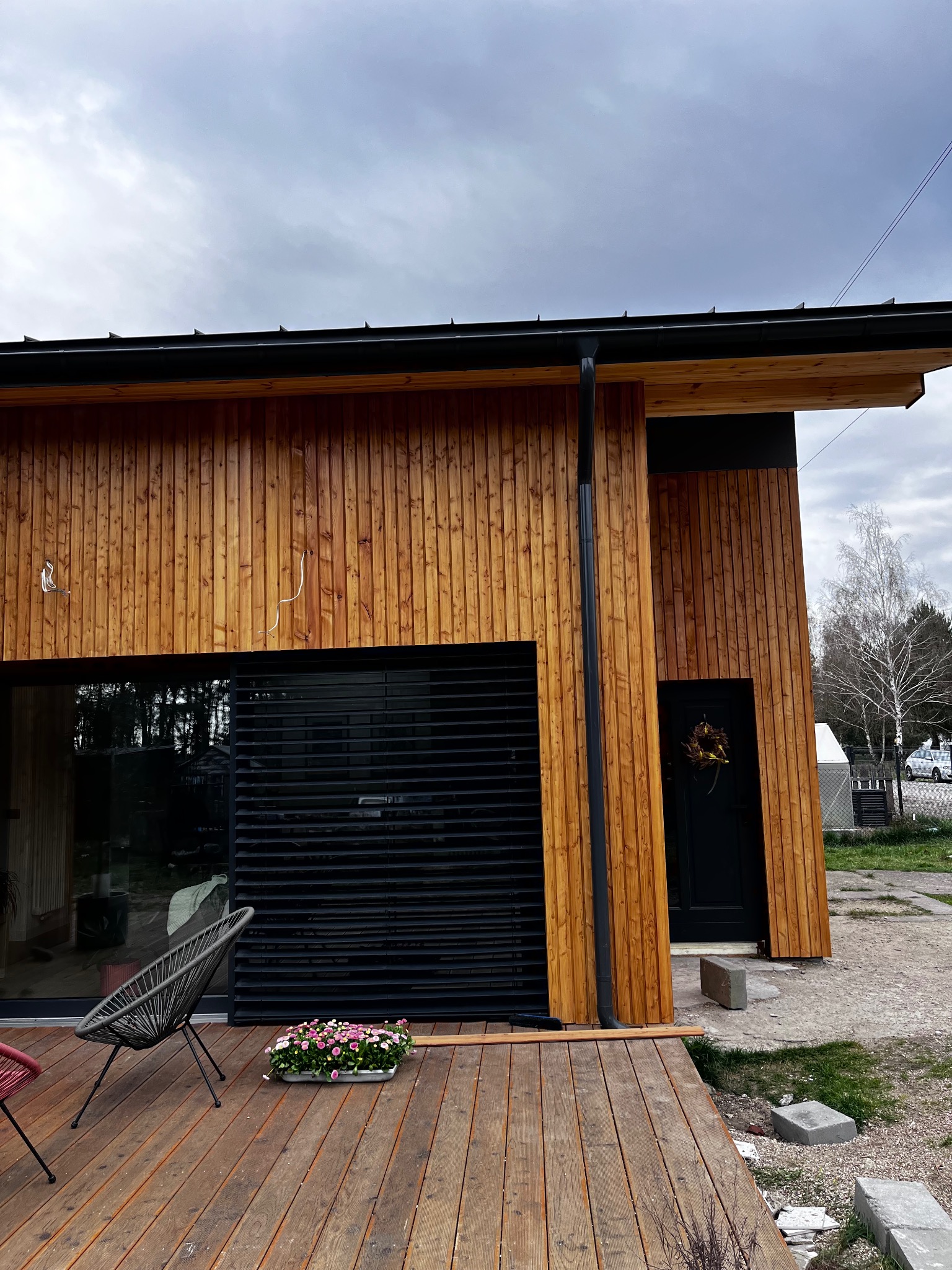 Nowoczesny dom z drewnianą elewacją, czarnymi żaluzjami i drewnianym tarasem. Widoczny fragment ogrodu i dekoracja na drzwiach wejściowych.