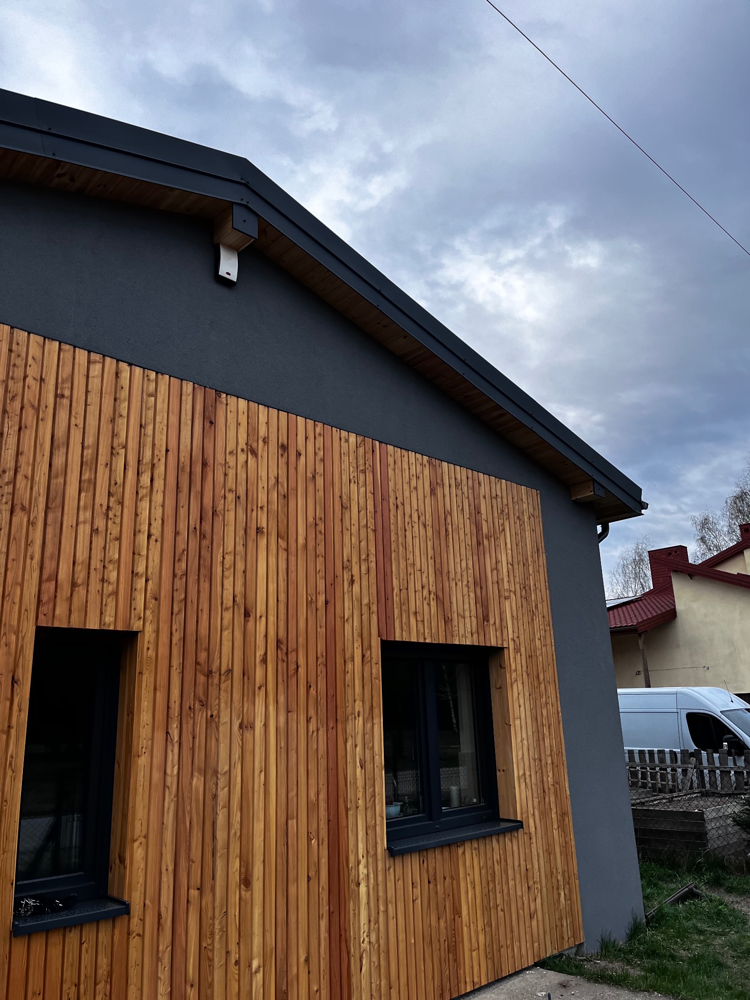 Elewacja domu z drewnianą okładziną i szarą strukturą, widok z dołu, z oknami w czarnych ramach. W tle budynki i błękitne niebo.