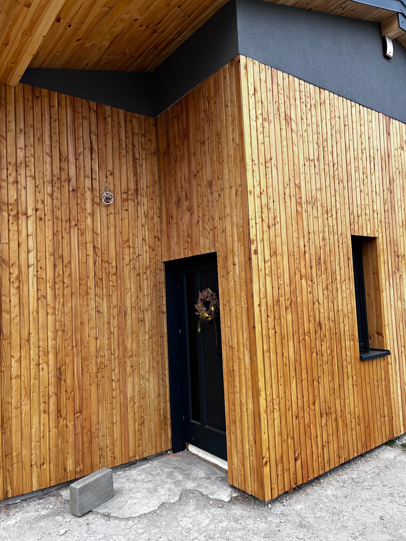 Elewacja domu z drewnianą okładziną pionową w ciepłym odcieniu brązu, czarne okna i drzwi. Widoczny fragment betonowej ścieżki i element dekoracyjny na drzwiach.