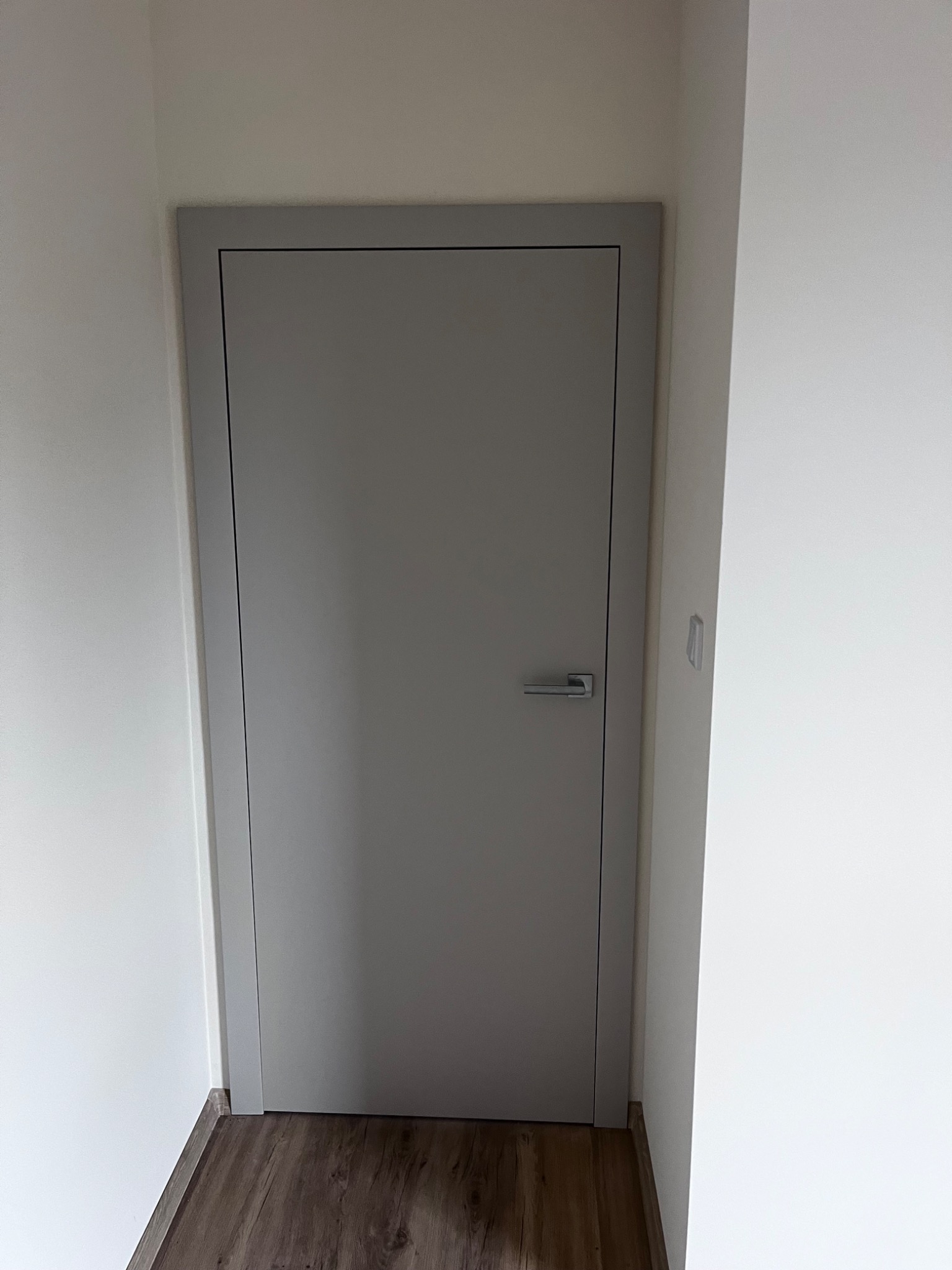 Zamknięte, gładkie, szare drzwi z srebrną klamką w białym wnętrzu. Widoczna podłoga z paneli imitujących drewno. Minimalistyczny design.