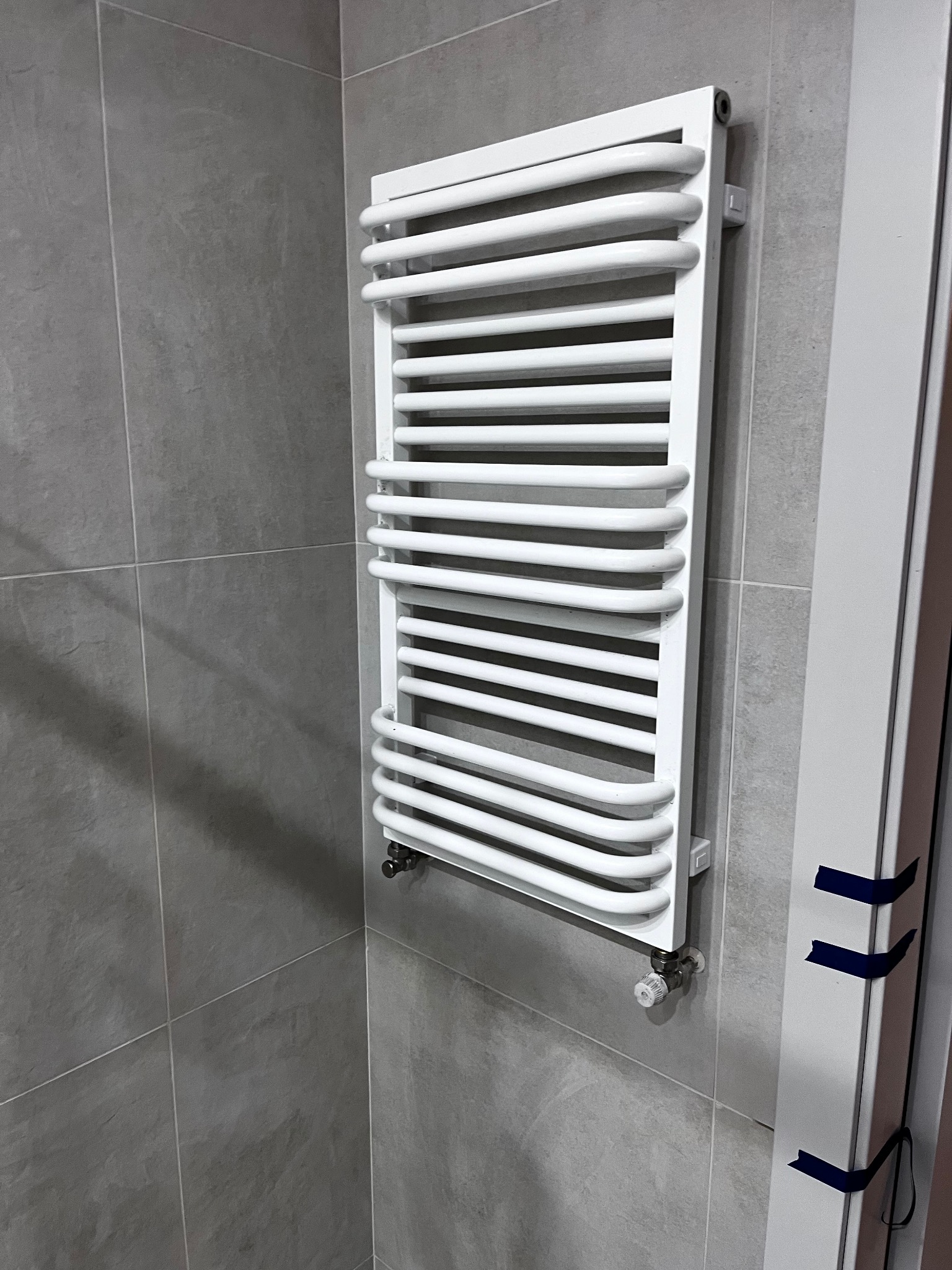 Biały, drabinkowy grzejnik łazienkowy na szarej, kwadratowej ścianie z płytek. Widoczne zawory i mocowania grzejnika. Fragment ościeżnicy drzwiowej z taśmą.