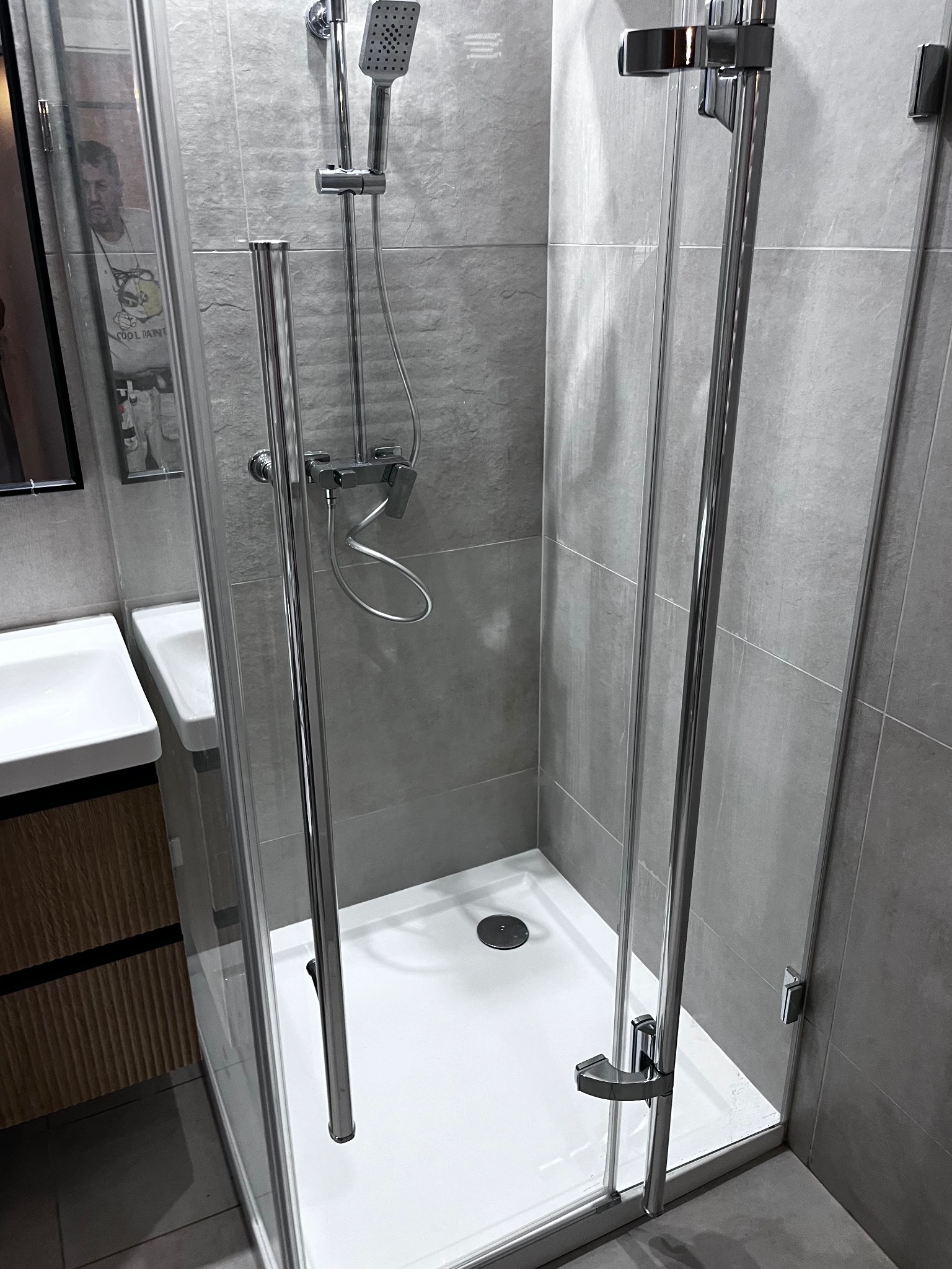 Nowoczesna kabina prysznicowa z chromowanymi detalami i szarymi płytkami. Widoczna umywalka i odbicie osoby w szkle. Minimalistyczny design.