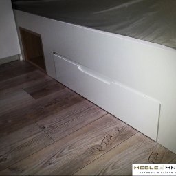 Meble Mnich - Białe łóżko z szufladami i otwartą półką, widoczne na tle podłogi z paneli. Minimalistyczny design, jasne kolory, drewnopodobne panele podłogowe.