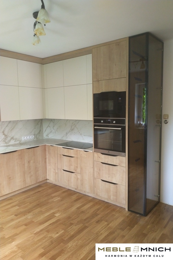 Jasna kuchnia na wymiar z drewnianymi frontami i marmurowym blatem. Zabudowa piekarnika i mikrofalówki, przeszklona szafka narożna.