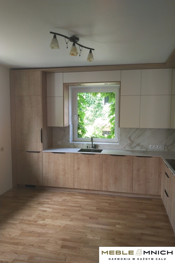 Jasna kuchnia z drewnianymi frontami i białymi szafkami wiszącymi, marmurowy blat i okno z widokiem na zieleń. Nowoczesne oświetlenie sufitowe.