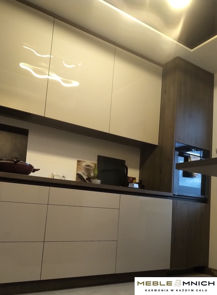 Nowoczesna kuchnia z białymi, lakierowanymi frontami szafek i drewnopodobnymi elementami wykończeniowymi. Widoczny ekspres do kawy i oświetlenie LED pod szafkami.