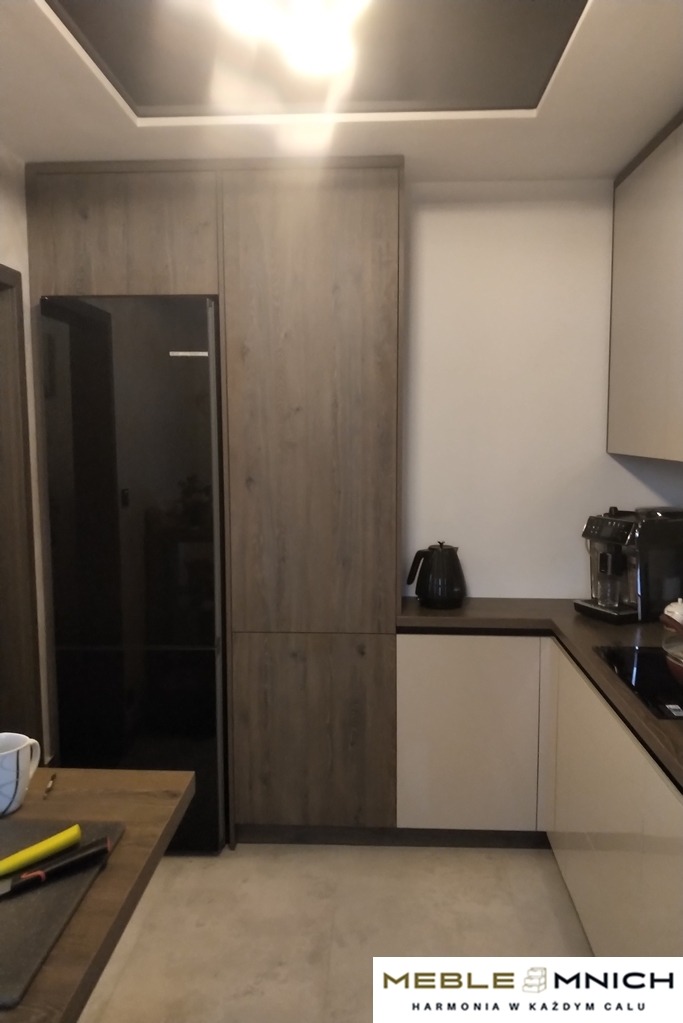 Nowoczesna kuchnia z zabudową w odcieniach brązu i bieli, z czarnym sprzętem AGD, oświetlona górnym światłem. Widoczny fragment blatu stołu.