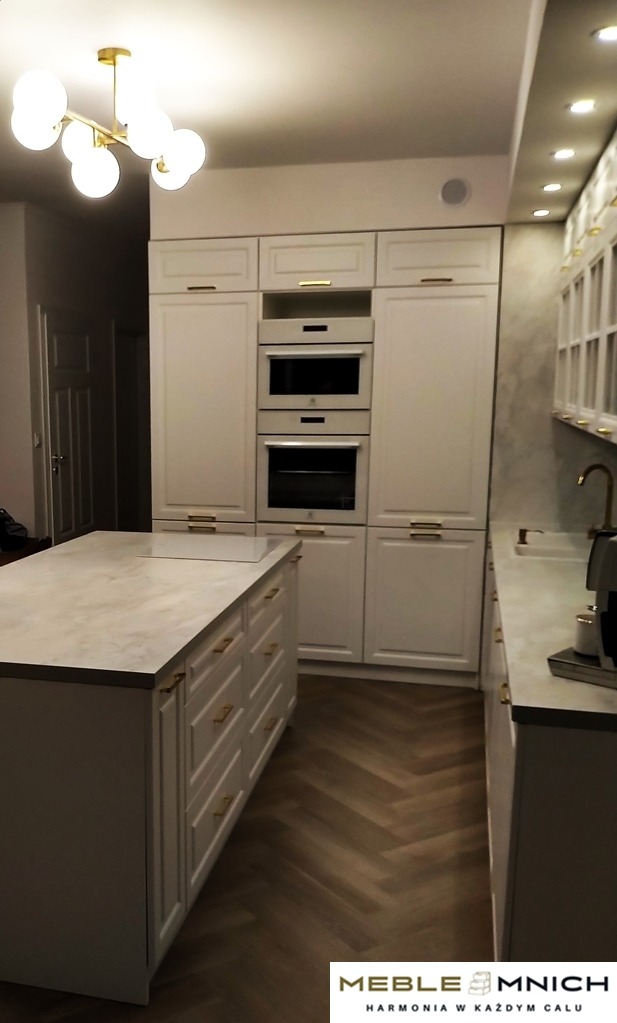 Elegancka, jasna kuchnia z wyspą i złotymi uchwytami. Parkiet ułożony w jodełkę. Nad wyspą widoczny nowoczesny żyrandol.