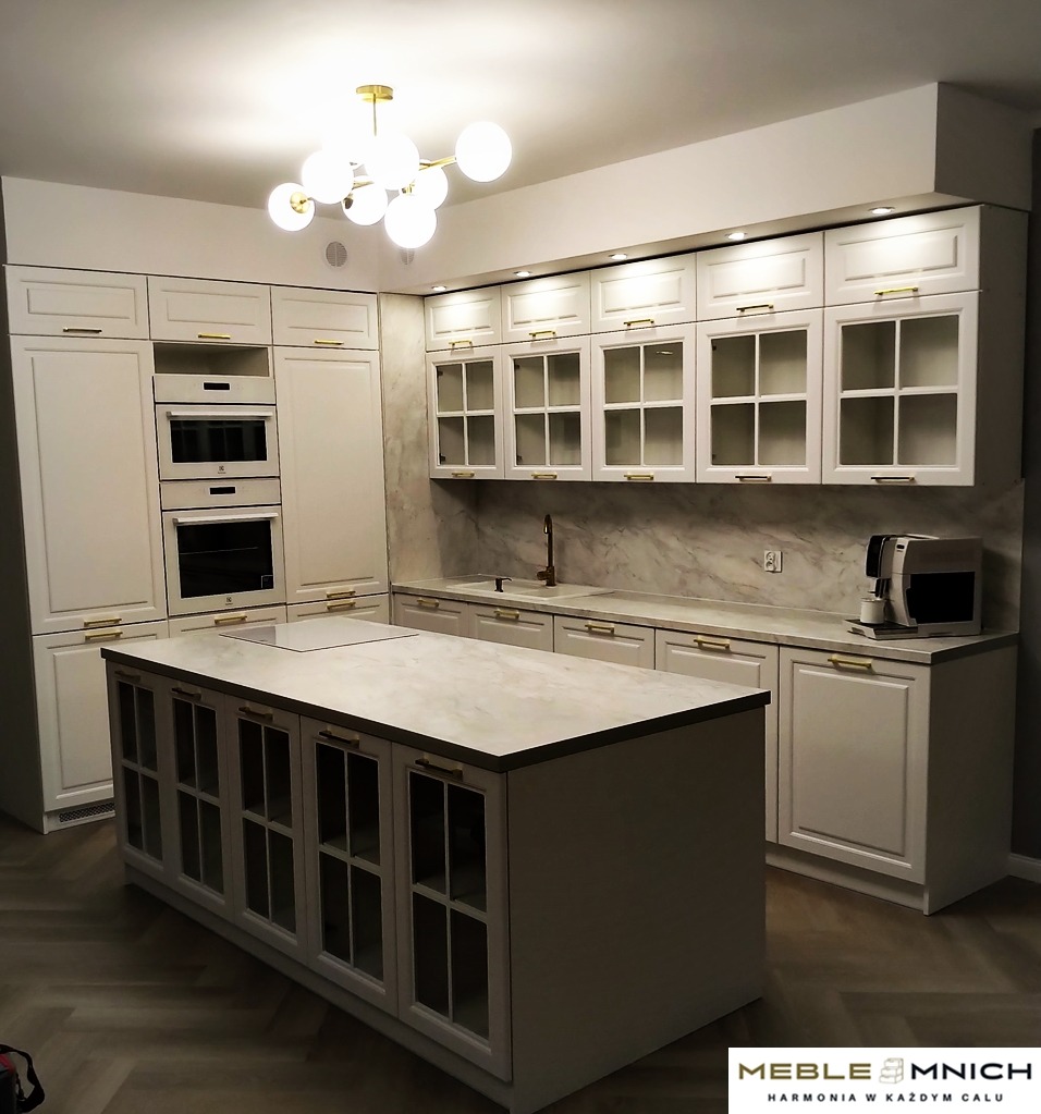 Jasna kuchnia na wymiar z wyspą i złotymi uchwytami. Górne szafki przeszklone, marmurowy backsplash, nowoczesna lampa sufitowa.