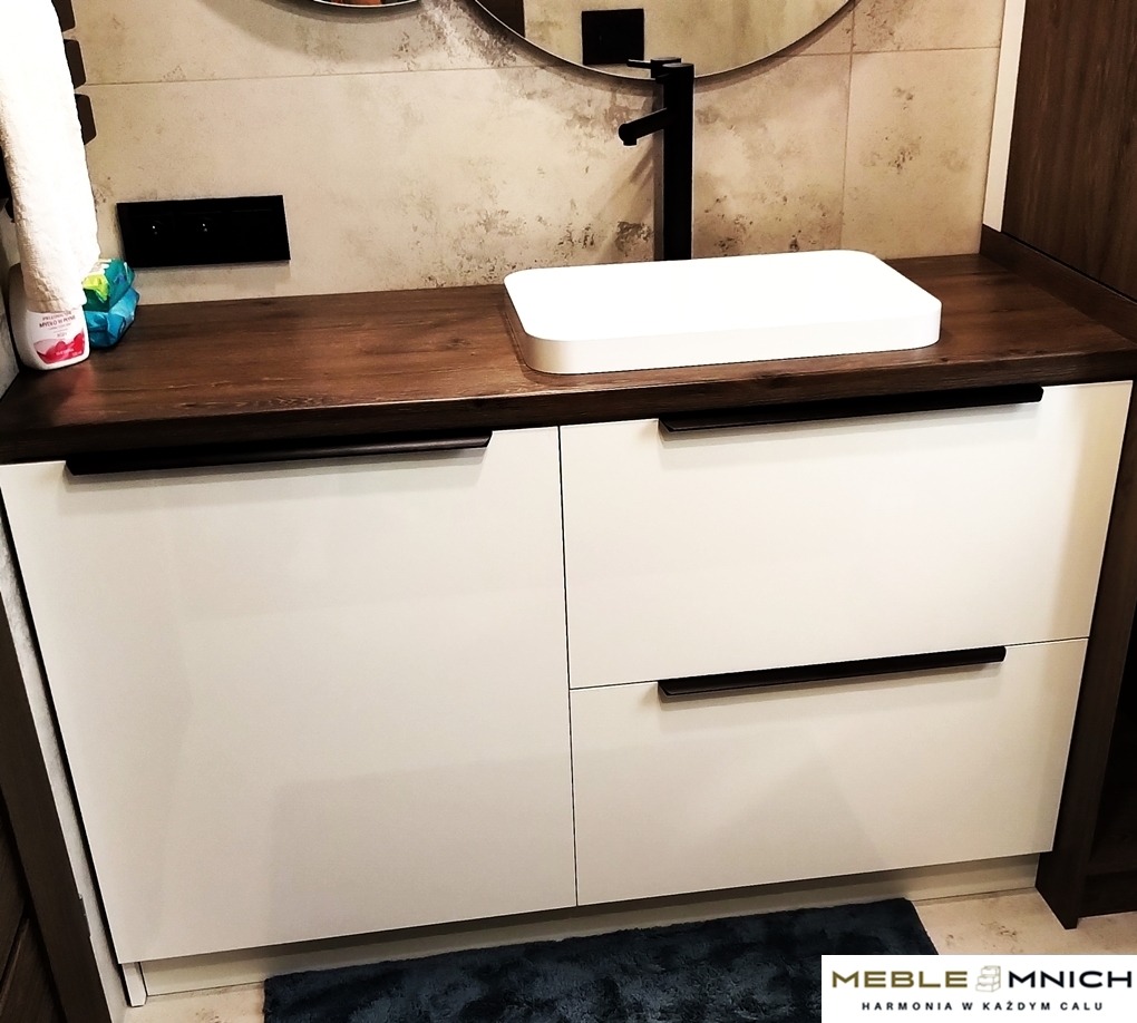 Biała szafka łazienkowa z blatem z ciemnego drewna i prostokątną białą umywalką nablatową, czarna armatura, okrągłe lustro w tle.
