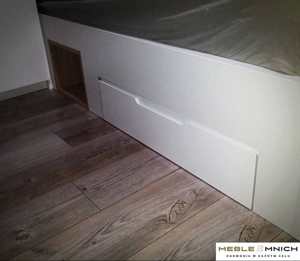 Białe łóżko z szufladami i otwartą półką, widoczne na tle podłogi z paneli. Minimalistyczny design, jasne kolory, drewnopodobne panele podłogowe.