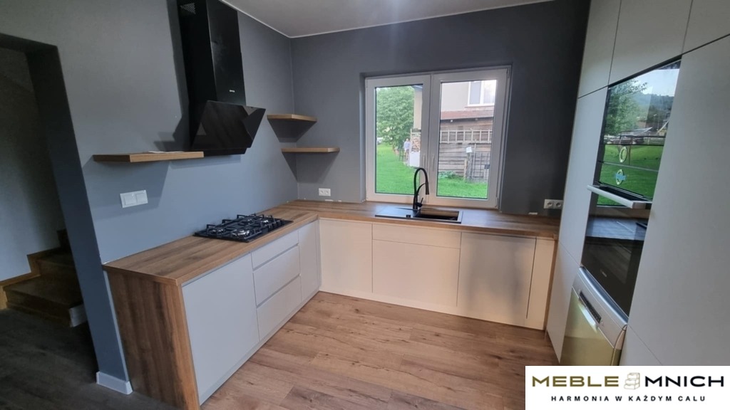 Jasna kuchnia w kształcie litery L z drewnianymi blatami i białymi frontami, czarny okap i płyta gazowa. Widok na zieleń za oknem i drewniane schody.