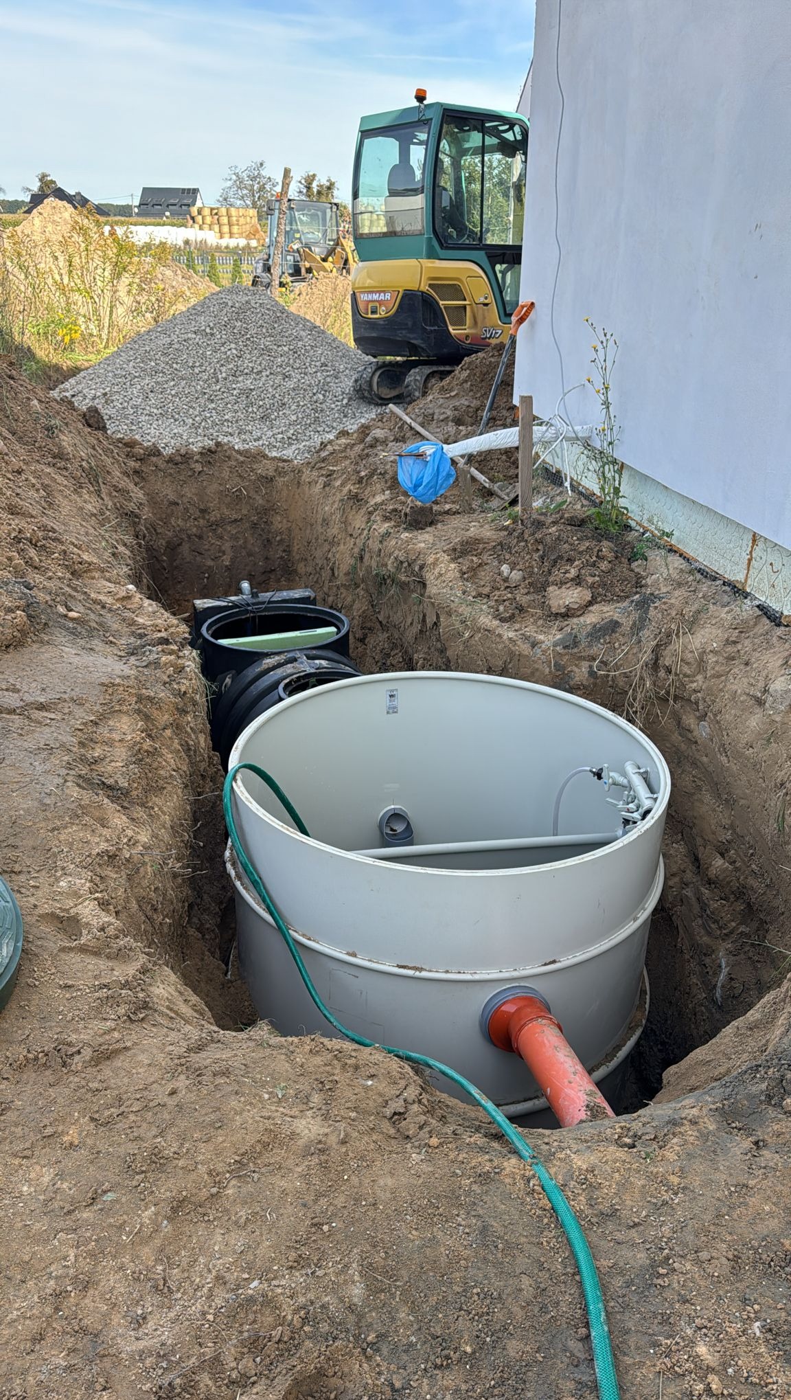 Szambo ekologiczne w wykopie obok budynku w Niechanowie. Widoczny mini-koparka i pryzma żwiru. Instalacja sanitarna z rurą odprowadzającą.