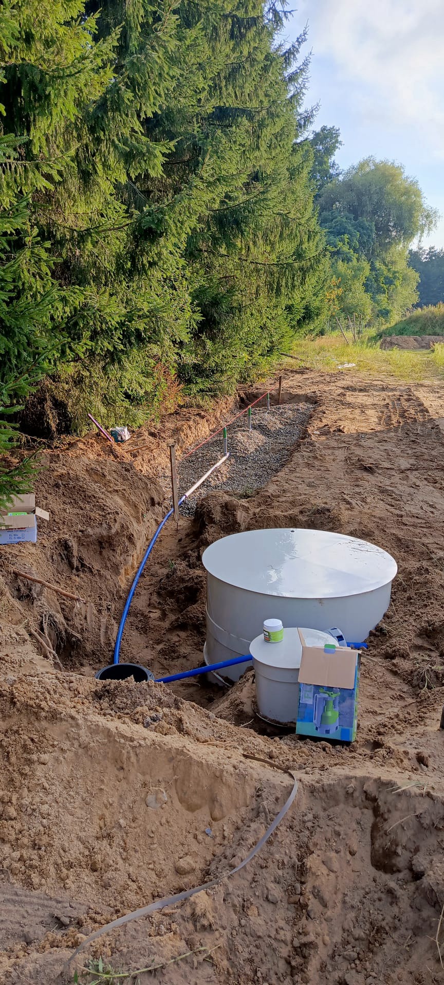 Biały zbiornik bezodpływowy w wykopie, niebieski wąż, obok pudełko z pompą. W tle przygotowany drenaż z kamieni i szpilki wyznaczające teren. Otoczenie: drzewa, ziemia.