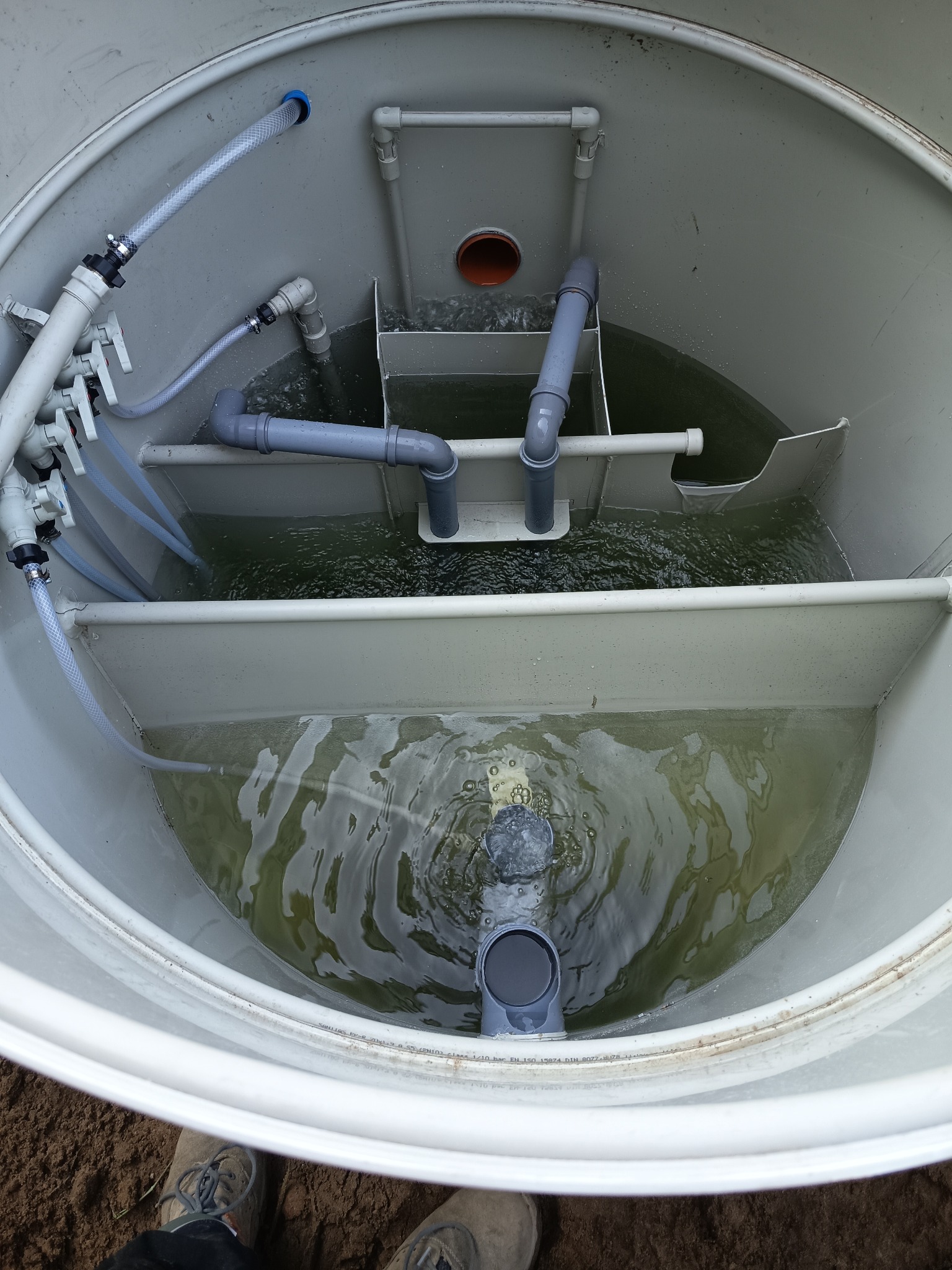 Wnętrze zbiornika z wodą i systemem rur PVC, prawdopodobnie szamba ekologicznego, widoczne z góry, z fragmentem butów w dolnym rogu kadru.