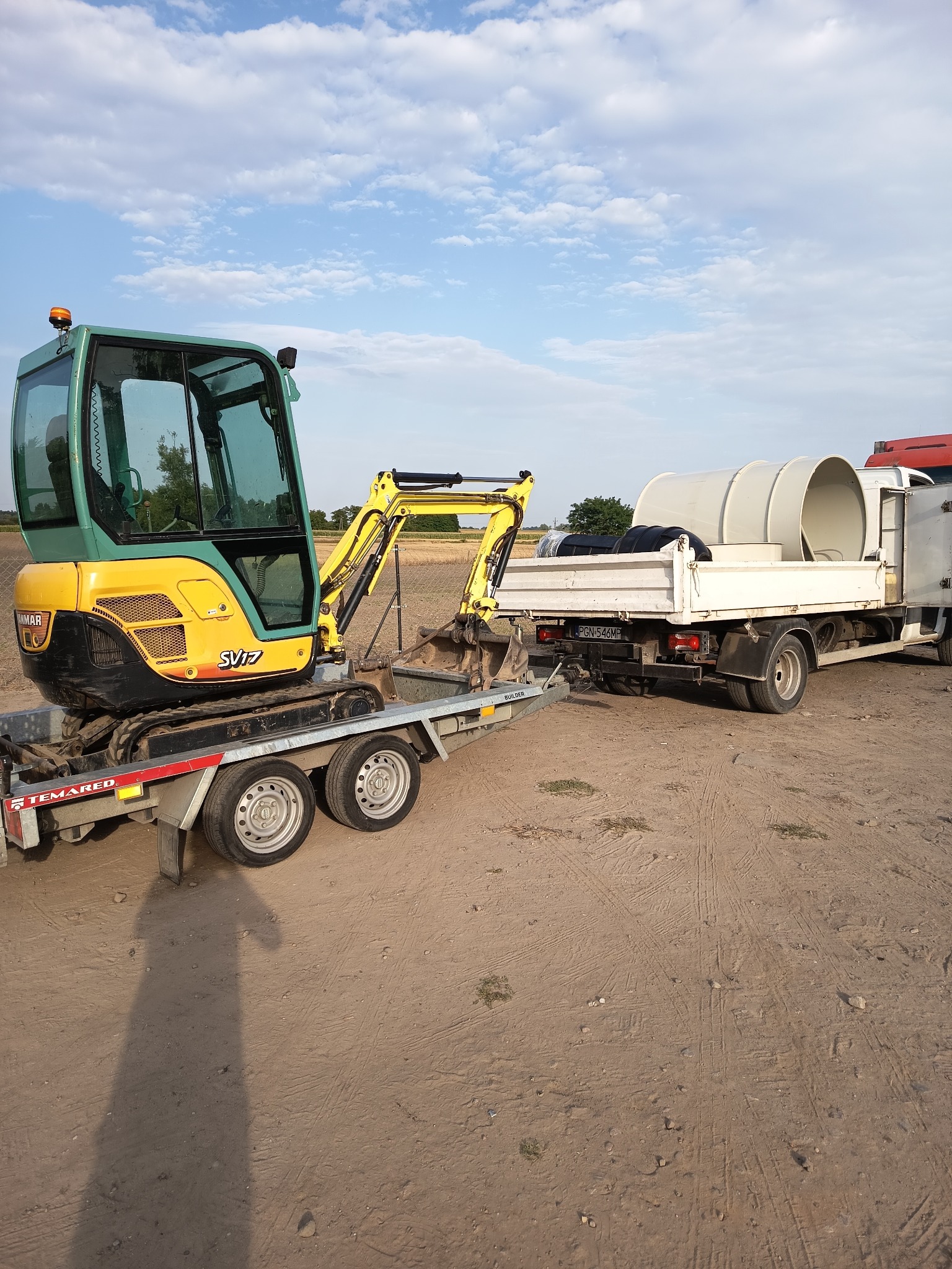 Żółto-zielona minikoparka Yanmar SV17 na przyczepie, obok ciężarówka z ładunkiem rur. Sprzęt na placu budowy, gotowy do prac ziemnych.