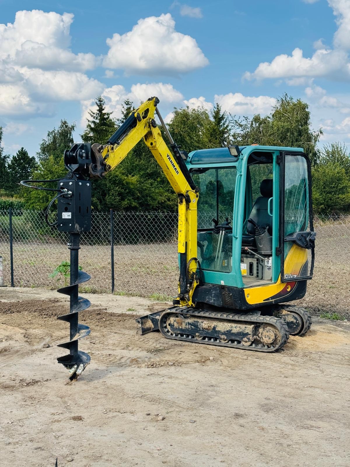 Żółto-turkusowa minikoparka Yanmar z wiertłem glebowym na placu budowy, widoczna świeżo wywiercona dziura w ziemi, w tle siatka ogrodzeniowa i drzewa.