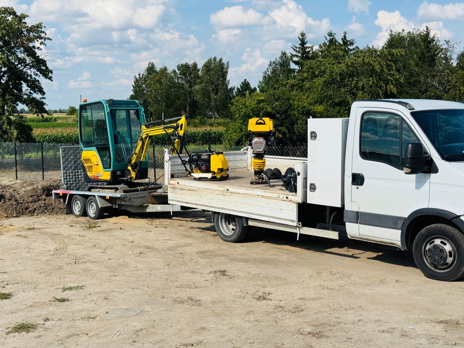 Żółto-zielona mini koparka na przyczepie załadowana na platformę samochodu dostawczego z ubijakami wibracyjnymi, na tle ogrodzenia i zieleni.