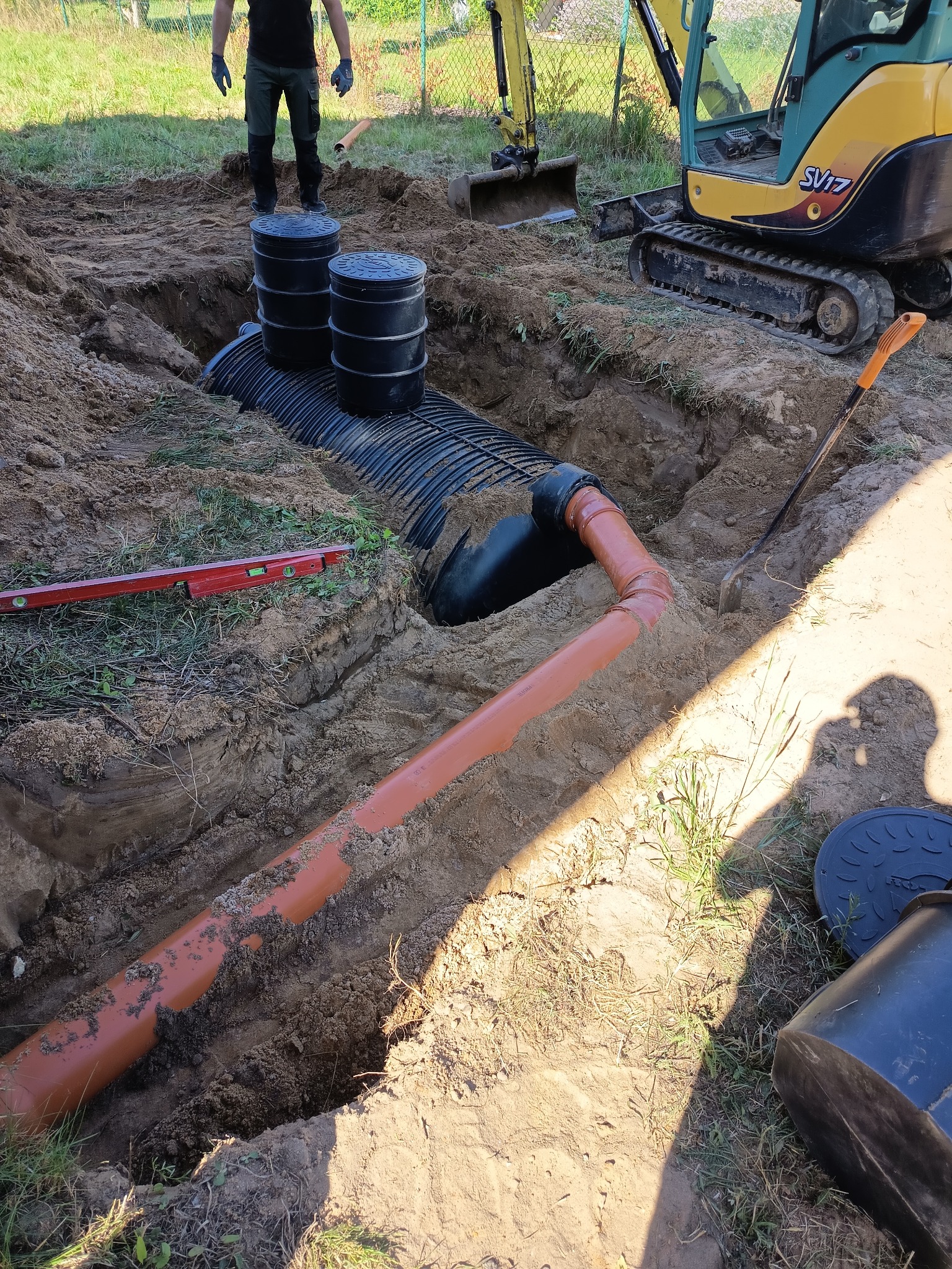 Wykop z ułożonymi rurami drenażowymi i studzienkami, widoczna koparka i poziomica. Prace ziemne w ogrodzie, częściowo zarośniętym trawą, w tle ogrodzenie z siatki.