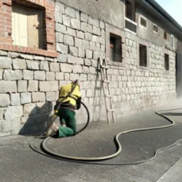 Pracownik w kombinezonie czyści kamienną elewację budynku metodą strumieniowo-ścierną. W tle widoczna drabina oparta o ścianę i okna z ceglaną obudową.