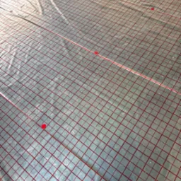 Mata grzewcza z czerwoną siatką i mocowaniami, przygotowana do instalacji ogrzewania podłogowego, widok z góry, jasne oświetlenie.