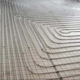 Białe rury ogrzewania podłogowego ułożone spiralnie na szarej macie z rastrem. Widok z góry, instalacja przed wylaniem posadzki. Równoległe ułożenie rur.