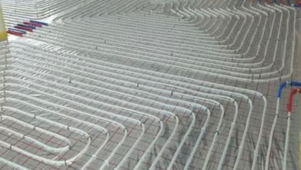 Ułożone białe rury ogrzewania podłogowego na szarej macie z siatką, widoczne niebieskie i czerwone złączki. Układ spiralny rur na dużej powierzchni.