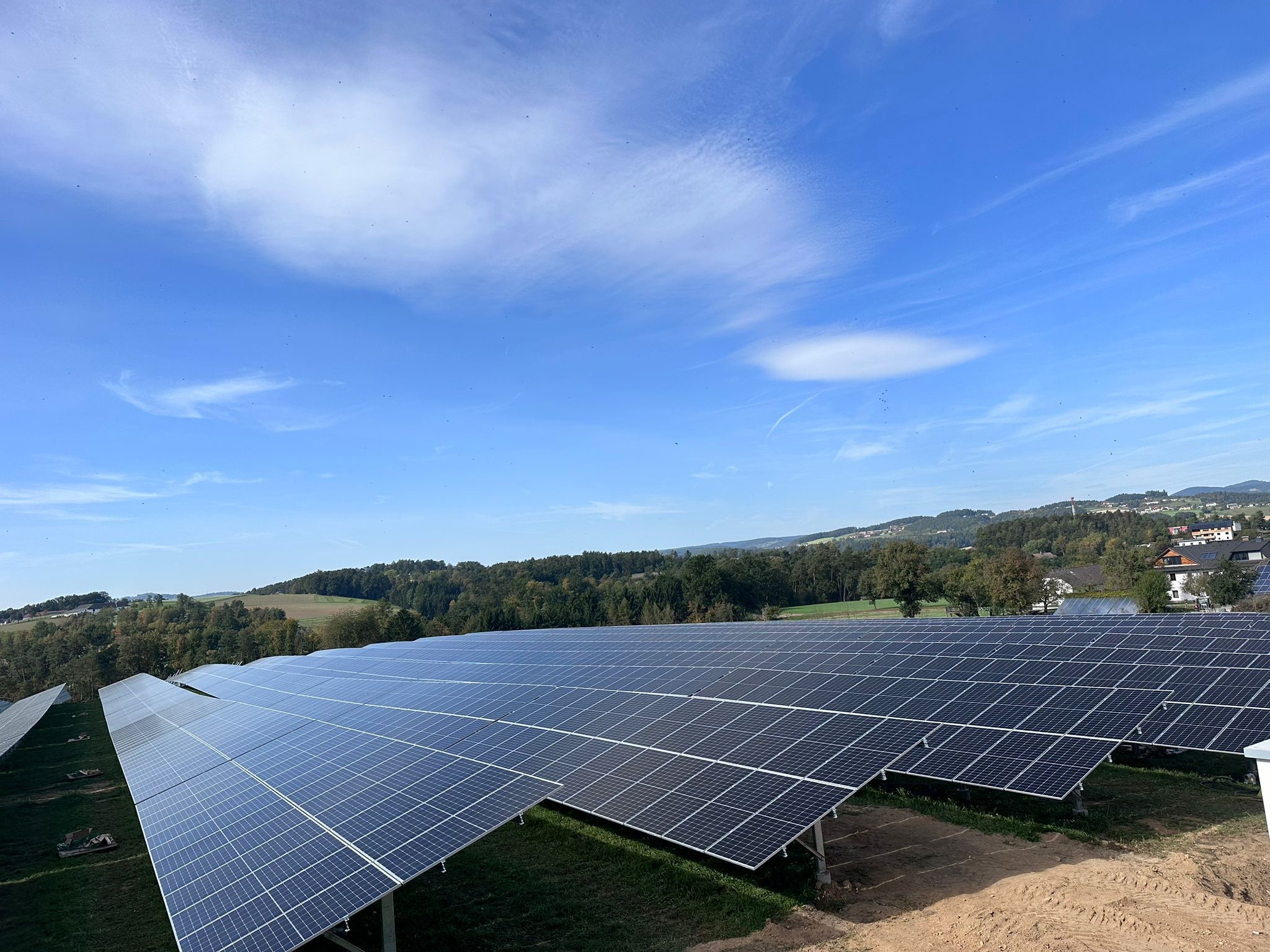 Rozległa farma paneli słonecznych na wzgórzu, błękitne niebo z chmurami w tle. Widok z góry ukazuje rzędy paneli i zielony krajobraz.