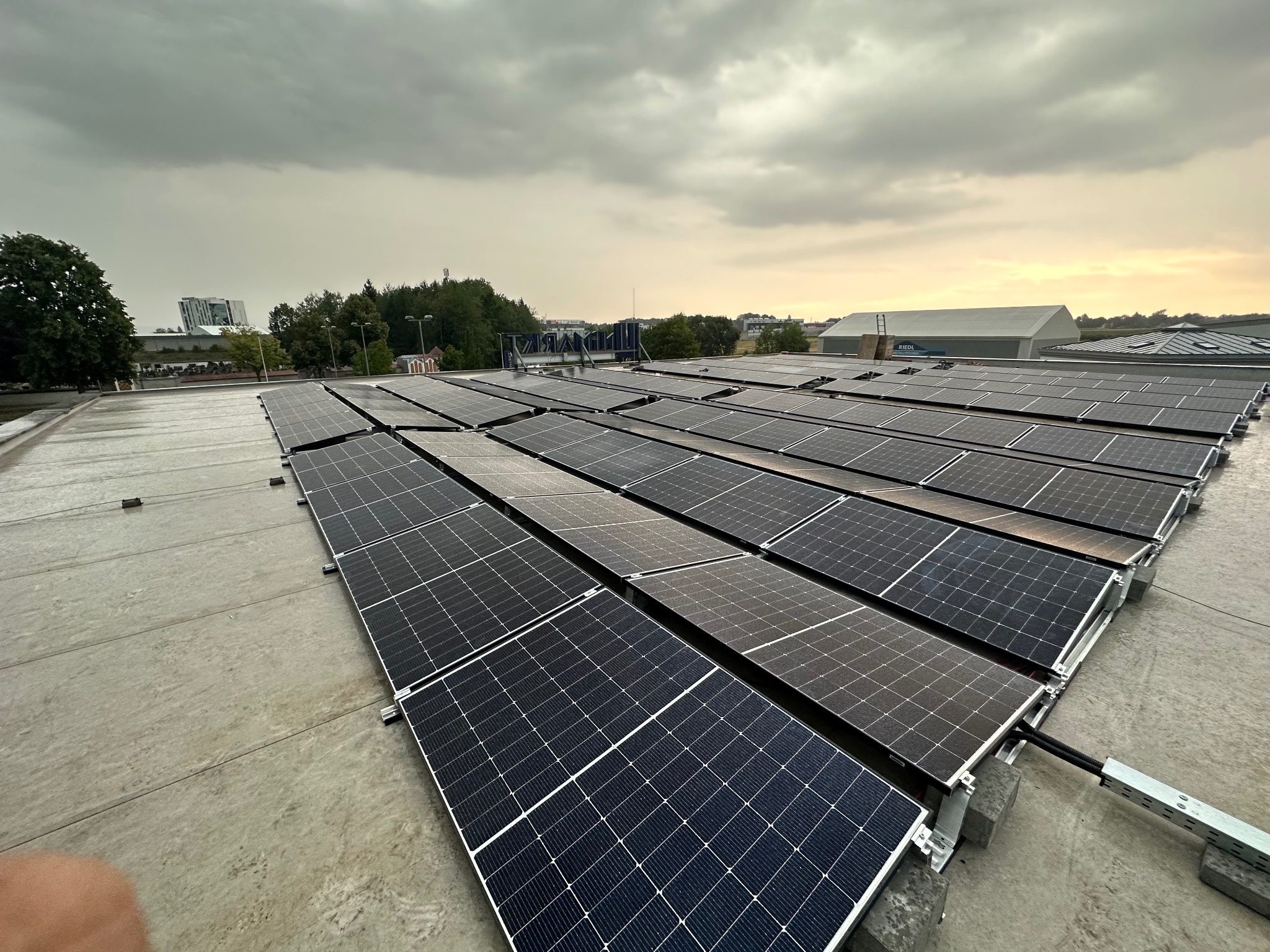 Rząd paneli słonecznych na płaskim dachu budynku w pochmurny dzień. Widoczne elementy montażowe i kable. W tle zabudowa miejska i drzewa.