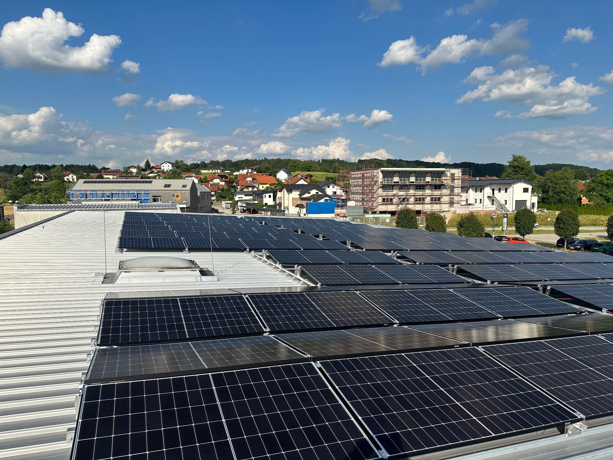 Instalacja fotowoltaiczna na dachu budynku z widokiem na okoliczną zabudowę i błękitne niebo z chmurami. Panele ułożone rzędami, odbijają światło.