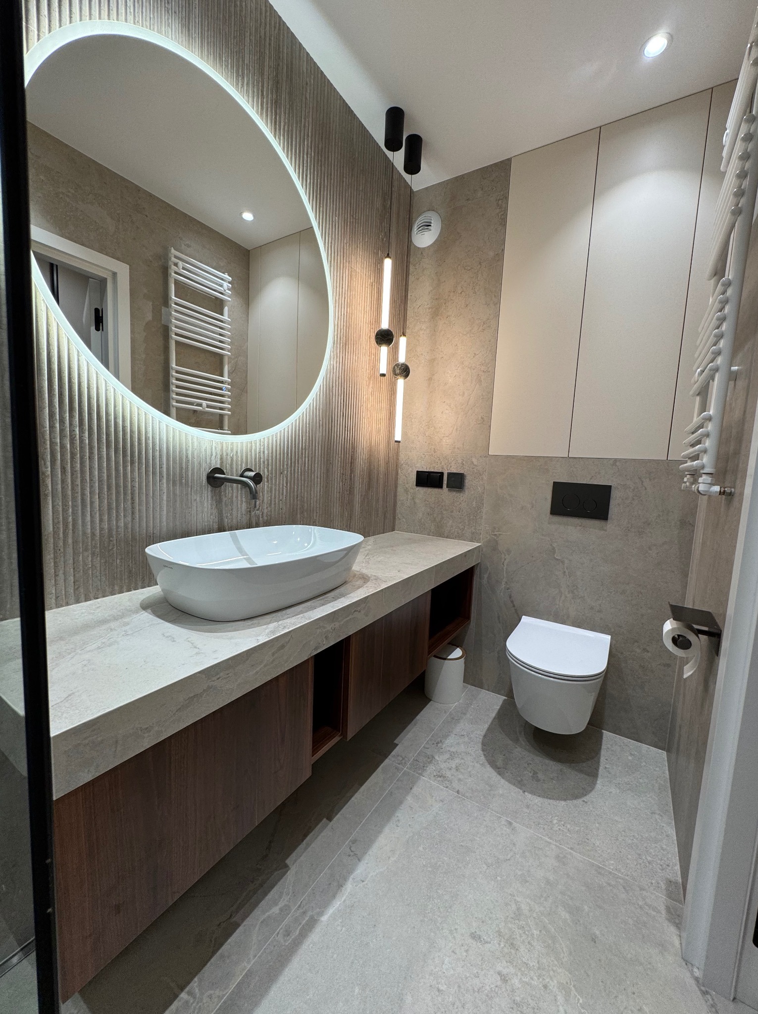 Nowoczesna łazienka z owalnym lustrem z podświetleniem LED, umywalką nablatową i wiszącą toaletą. Beżowe płytki imitujące kamień i drewniane akcenty.