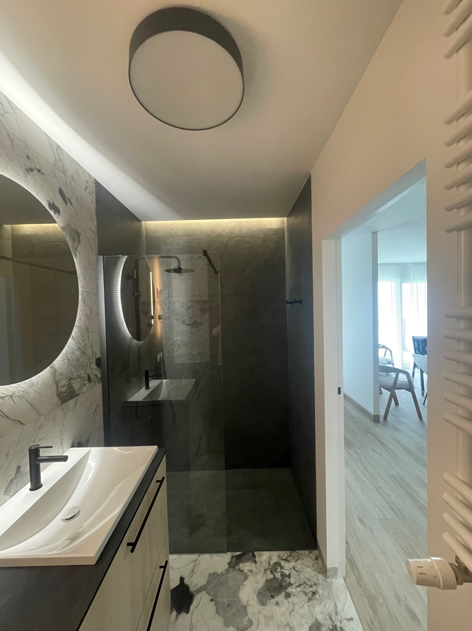 Nowoczesna łazienka z kabiną prysznicową typu walk-in, okrągłym lustrem z podświetleniem LED i marmurową posadzką. Minimalistyczny design, czarne akcenty.