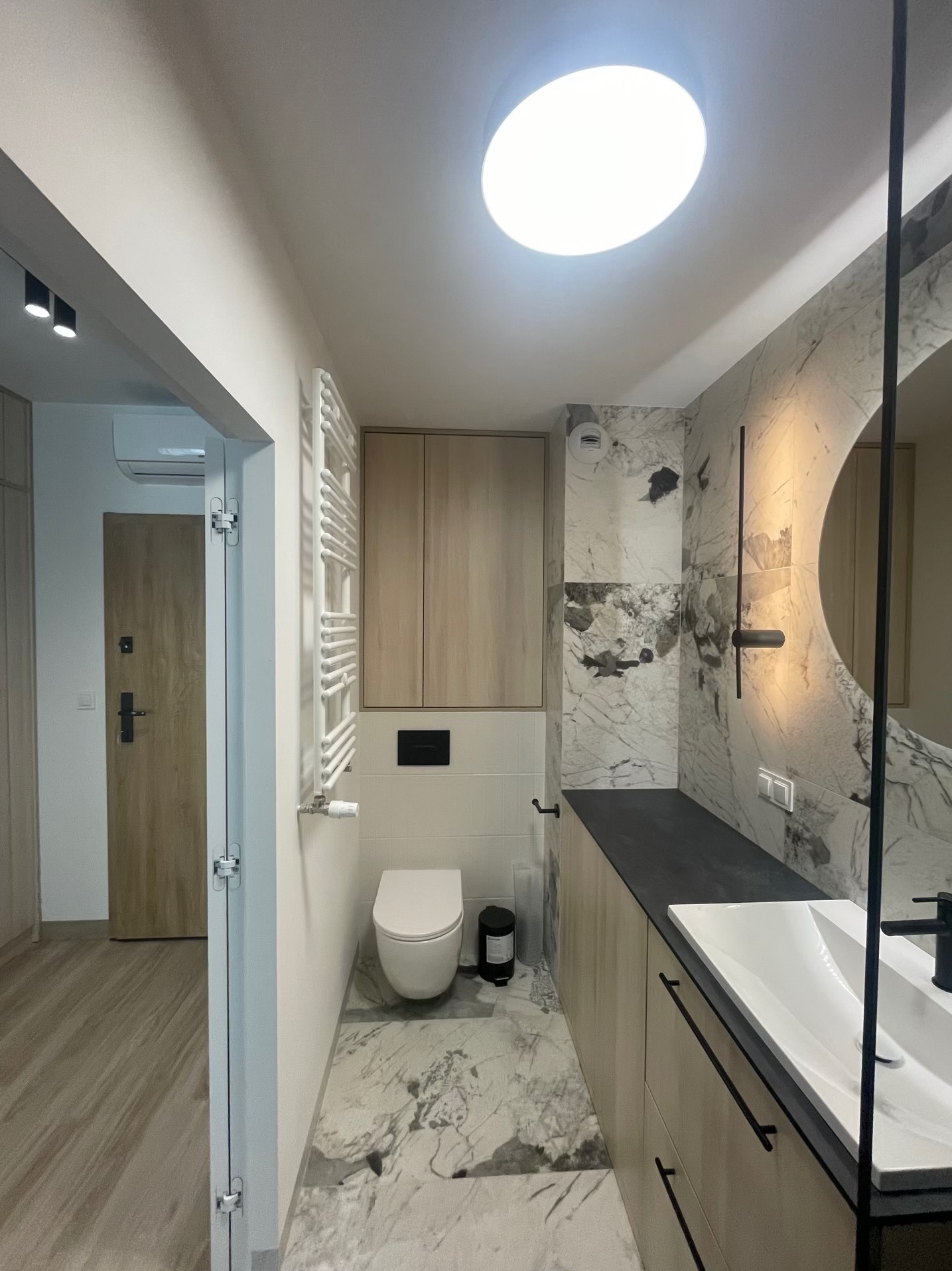 Nowoczesna łazienka z marmurowymi płytkami, drewnianymi szafkami i okrągłym lustrem. Widoczny grzejnik drabinkowy i oświetlenie sufitowe.