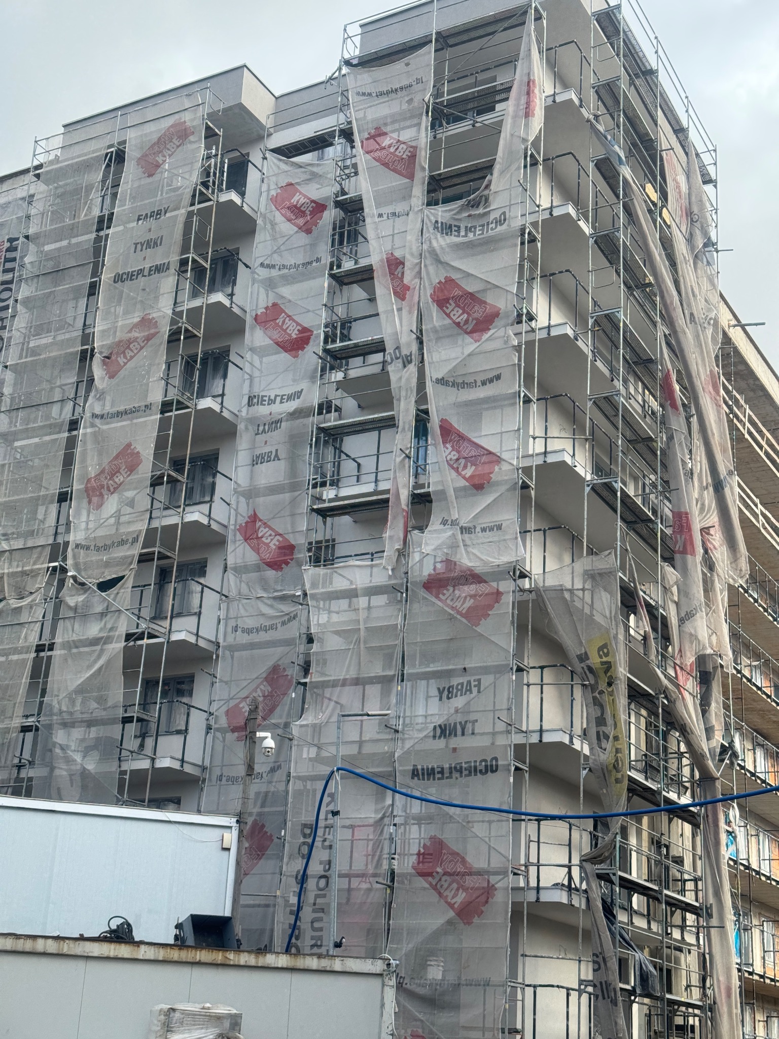 Budynek w rusztowaniach, osłonięty siatką z logo producenta farb i tynków. Prace elewacyjne w toku, widoczna instalacja zabezpieczająca.